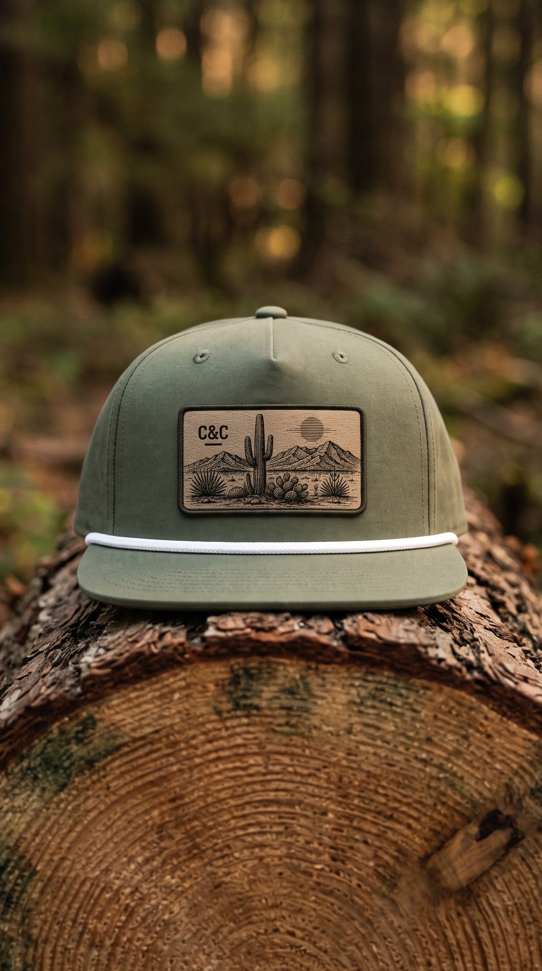 An olive green flat-brim baseball cap with a rectangular desert landscape patch on the front, resting on a cut log in a forest setting.